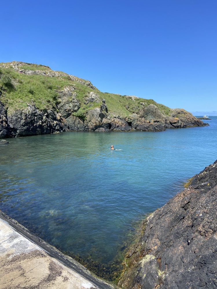 an image showing green headland, blue sky and blue-green sea with a woman swimming.
