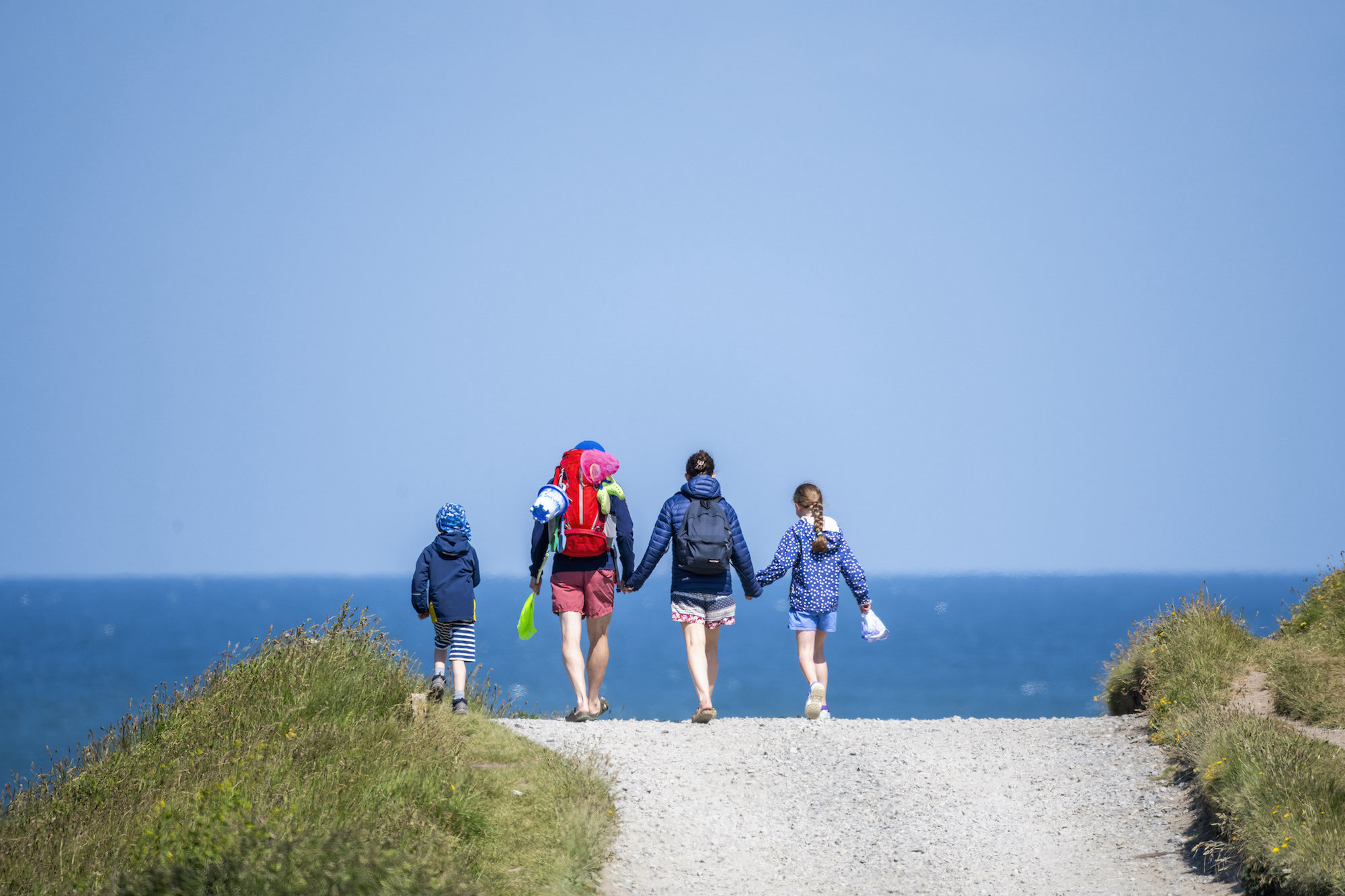 Family holding hands walking towards blue sea on coastal path in Nefyn, North Wales
