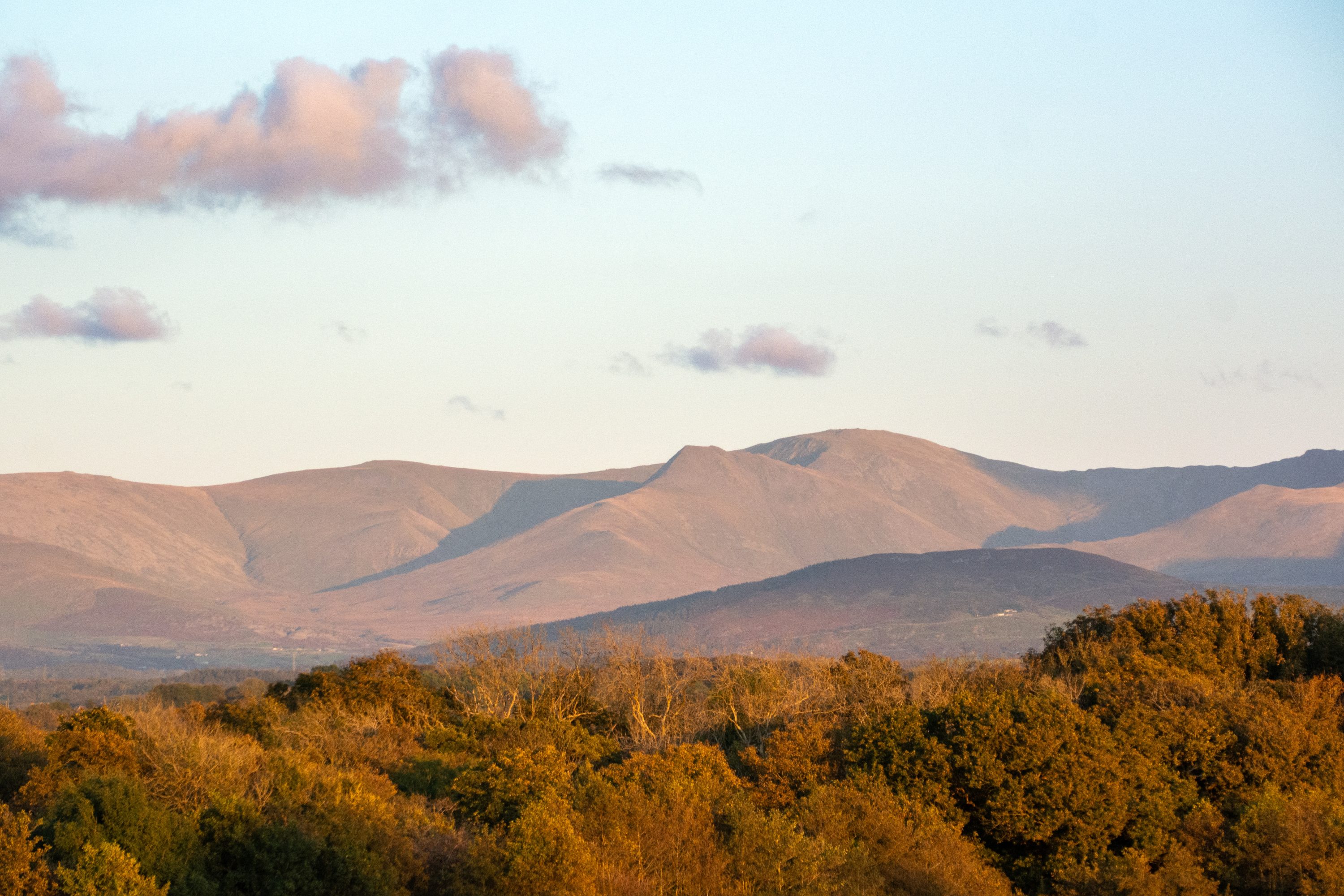 Sunset with blue skies and pink clouds behind Snowdonia (Eryri) mountain range with warm coloured Autumn trees in the foreground.