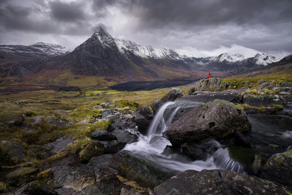 A moody image of the mountains and lakes in Snowdonia North Wales with a walker in a bright red coat enjoying the view.